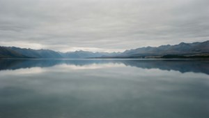 A calm lake reflects surrounding mountains under an overcast sky. — Grey Picture
