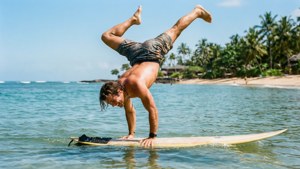 Leikkisä surffaaja tekee käsilläseisontaa surffilaudalla kristallinkirkkaassa trooppisessa vedessä.