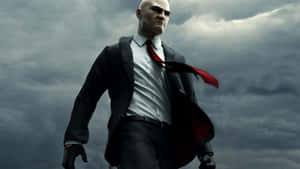 A bald assassin in a black suit and red tie walks under a stormy sky.