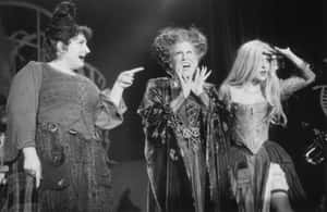 Tres brujas teatrales disfrazadas gesticulando en un escenario oscuro durante una actuación en vivo. — Estética Hocus Pocus Imagen Fondos de pantalla