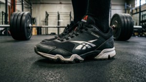 Foto sudut rendah sepatu Reebok CrossFit di gym dengan barbel.
