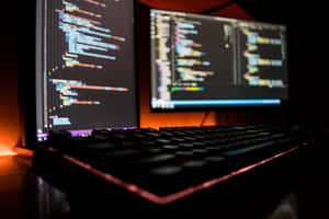 Two computer monitors showing code with a glowing keyboard in a dark room. — It Computer Picture