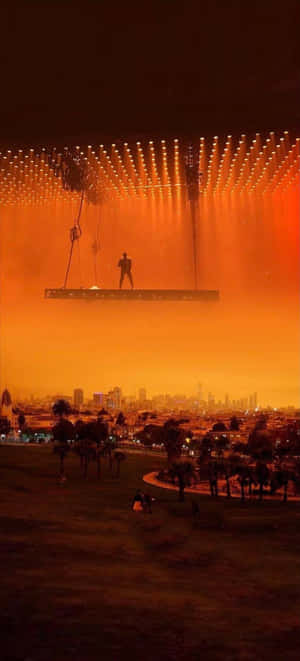 Um artista em um palco flutuante iluminado por luzes laranja sobre uma cidade à noite. — Kanye iPhone Papéis de Parede