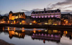 Edifícios históricos ao longo de um rio ao entardecer com luzes coloridas refletindo na água. — Laval Papéis de Parede