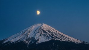 Uma vista telefoto de um pico de montanha nevado com uma lua crescente em um céu crepuscular.
