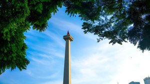 Tall gray monument with red flag framed by green trees against blue sky. — Jakarta Background