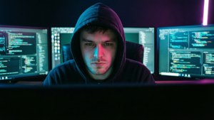 Eine vermummte Gestalt, die an mehreren Monitoren mit Neonbeleuchtung codiert
