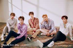 Five young men in stylish outfits sitting on a patterned rug in a bright studio. — Nuest Picture