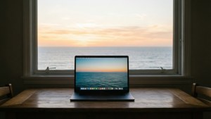 En bærbar computer ved vinduet med havudsigt ved solopgang.