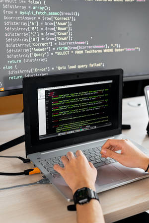 Person coding on laptop with monitor showing PHP code in a tech workspace. — Computer Screen Wallpaper