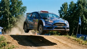 La Ford Fiesta RS di Ken Block prende aria sopra una cresta di rally, incorniciata da alberi e cielo blu.