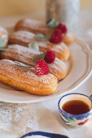 Fila di eclair con zucchero a velo guarniti con lamponi e foglie di salvia accanto a una tazza di tè floreale.