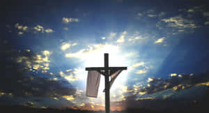 Silhouette of a cross with a cloth draped over it against a sunset sky with radiant sun rays.