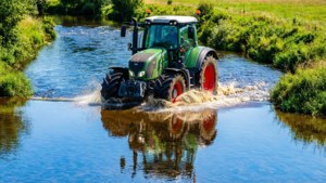 Een blauwe Fendt-tractor die een ondiepe rivier oversteekt met spatten.