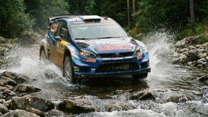 Rallyauto ylittää matalan joen, ja vesi roiskuu renkaiden ympärillä.