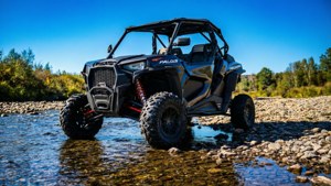 RZR XP 1000 aseteltuna märillä kivillä joen varrella, eloisa sininen taivas ja veden heijastukset