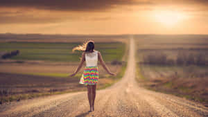 Woman walking barefoot on a dirt road at sunset with arms outstretched. — Road Wallpaper
