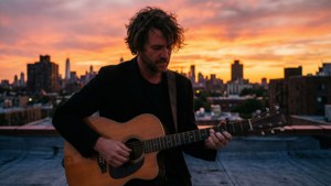 Beck spiller guitar på en tagterrasse under solnedgang med byens skyline