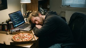 Müder Programmierer schläft an einem unordentlichen Schreibtisch mit Energy-Drinks und Pizza