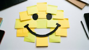 Wallpaper flat-lay yang ceria dari catatan Post-it yang disusun menjadi wajah smiley di atas meja putih.