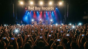 Perspektif panggung dari kerumunan konser besar dengan spanduk Bad Boy Records, lampu biru dan merah, energi mentah.
