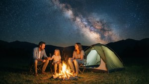 Uma família acampando em uma barraca sob um céu estrelado com a Via Láctea visível, fogueira brilhando suavemente.
