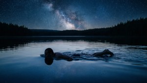 Person floating on a calm lake under a starry night sky with Milky Way visible.