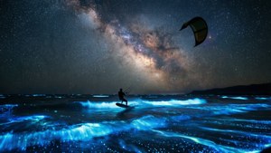 Kitesurfer sob a Via Láctea com ondas bioluminescentes.