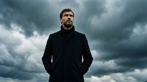 Episches Weitwinkelporträt von Pep Guardiola, der in einem schwarzen Mantel zu einem dramatischen bewölkten Himmel aufblickt