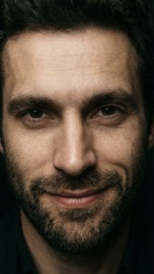 Primo piano intenso del volto di Vincenzo con un leggero sorrisetto, alto contrasto e sfondo scuro.