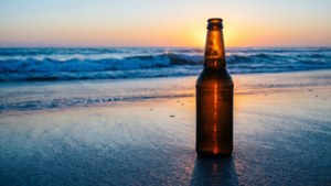 Uma garrafa de cerveja repousa em uma praia de areia enquanto o sol nasce sobre ondas calmas.