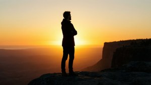 Silhouette einer Figur auf einer Klippe bei Sonnenuntergang, Arme verschränkt, vom Himmel abgewandt.