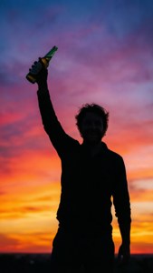 Silhueta de uma pessoa brindando com uma garrafa de cerveja contra um pôr do sol colorido.