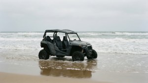 Een grijze buggy rijdt door de branding op een eenzaam strand onder een bewolkte lucht.
