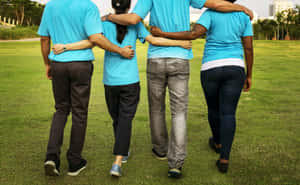 Four people walking together arm-in-arm on a grassy field wearing blue shirts. — Teams Background
