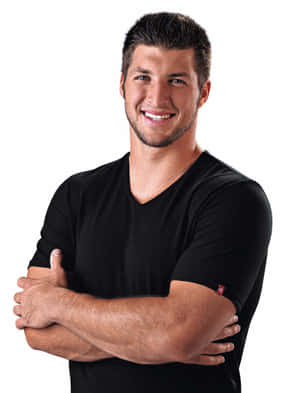 A smiling man in a black t-shirt with arms crossed in a studio portrait. — Tim Tebow Picture