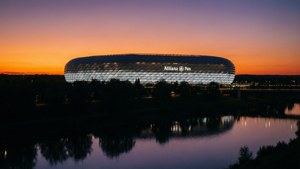 Allianz Arena i skymningen reflekterad i en lugn flod med orange-lila himmel