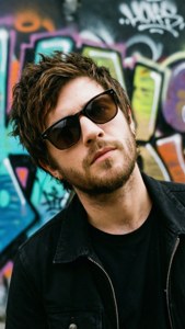Niall Horan in un ambiente urbano con muro di graffiti, occhiali da sole e un'espressione cool.