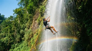 Wallpaper yang hidup dan dinamis dari seorang pemanjat yang rappel menuruni air terjun dengan pelangi di semprotan air.